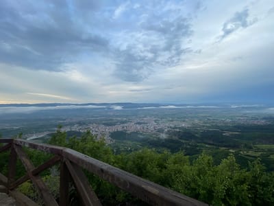 The image is a high-angle view overlooking the city of Delchevo, Bulgaria, under a cloudy sky. In the foreground, a wooden railing, weathered and marked with graffiti, runs horizontally across the frame. Dense green foliage, primarily trees with sparse white blossoms, surrounds the railing.
The middle ground reveals the city of Delchevo spread out below, characterized by a dense cluster of buildings with red-tiled roofs. The city appears nestled within a valley, with patches of greenery and roads weaving through it. Some areas exhibit a slight haze, possibly indicating mist or atmospheric conditions.
The background features a vast, undulating landscape of hills and mountains stretching towards the horizon. The sky is filled with dramatic, layered clouds, ranging from light grey to darker hues, suggesting an overcast or approaching weather system. The overall lighting is diffused, contributing to a muted color palette. There are no people or animals visible in the image. The scene appears to be captured from a vantage point, likely a scenic overlook or viewpoint, given the railing and the expansive panorama.