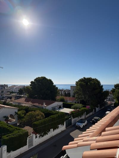 The image captures a bright, sunny day in Alcalà de Xivert, Spain, likely from an elevated vantage point, possibly a rooftop or balcony. The foreground is dominated by the terracotta tiles of a roof, sloping downwards towards the right. Beyond the roof, a street lined with white-walled houses and manicured green hedges is visible. Two cars are parked on the street: a white crossover vehicle and a dark-colored SUV. Further in the distance, a line of buildings, some with tiled roofs, are situated before the shimmering blue Mediterranean Sea, which stretches to the horizon under a clear, bright blue sky. The sun is prominently visible in the upper left quadrant of the sky, casting a strong glare and lens flare. A faint contrail from an aircraft is visible in the sky. The overall scene depicts a residential area by the coast on a clear, warm day.