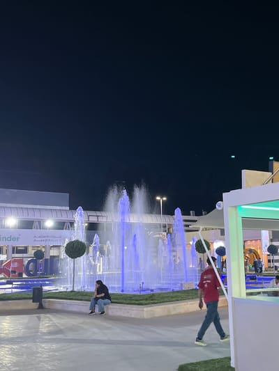 An outdoor fountain plaza is captured at night in Al-Ghizlaniya, Syria. The fountain, illuminated with vibrant blue lights, is the central focus, with water jets reaching high into the dark sky. In the foreground, a man in a red shirt and blue jeans walks away from the camera. To the left of the fountain, a woman with dark hair sits on a stone ledge, looking to the side. Behind the fountain, a building facade is visible with Arabic text, indicating a commercial or business area. The overall scene depicts a lively public space at night, with people enjoying the illuminated fountain.
