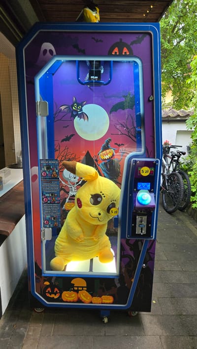 In the city of Łagów, Poland, an outdoor claw machine, decorated with a Halloween theme featuring ghosts, bats, a skeleton, and pumpkins, is positioned on a paved walkway. The machine's glass front showcases a large, yellow Pikachu plush toy as the prize. A sign inside the machine, written in Polish, provides instructions for use. To the right of the machine, two bicycles are parked, and behind them, a building with greenery is visible. The overall scene depicts a recreational area possibly near a park or a commercial establishment.