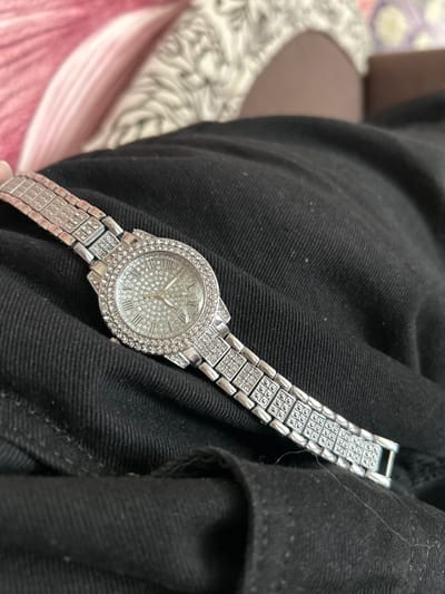 In Dmitrievskaya, Russia, a silver-toned wrist watch is depicted resting on a dark, folded textile surface consistent with black denim. The watch features a round dial entirely covered in small, clear, faceted stones, displaying silver-toned Roman numeral hour markers (I, II, IV, V, VII, VIII, X, XI, XII are discernible) and three slender hands. The watch bezel is also embellished with two concentric rows of similar clear, faceted stones. The metallic band is comprised of multiple rectangular links, each segment containing a grid of clear, faceted stones. The background is out of focus, revealing blurred patterns of pink fabric in the upper left and a light-colored, textured fabric adjacent to a brown surface in the upper right. No individuals or active interactions are present in the scene.