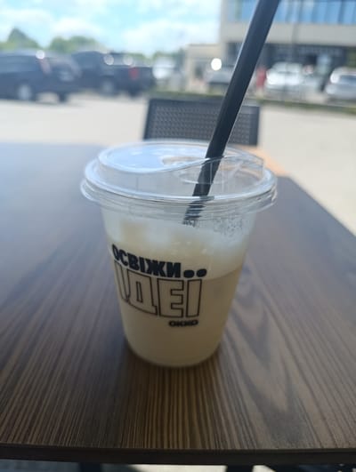 A plastic cup with a clear lid and a black straw sits on a wooden table. The cup contains a light-colored beverage with foam on top. Text is visible on the cup, appearing to be in Ukrainian. In the background, out of focus, are parked vehicles, trees, and a building under a cloudy sky. The scene suggests an outdoor seating area, possibly at a cafe or restaurant, in Berdychiv, Ukraine. There are no people visible in the image.
