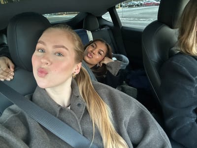 The scene depicts three individuals inside a dark-colored vehicle, likely a Tesla, in Solna, Sweden. In the foreground, a young adult female with light blonde hair pulled back in a ponytail, wearing a grey coat and a seatbelt, faces the camera. She has blue eyes, gold hoop earrings, and is making a "kiss" face with pursed lips. Her left hand, with gold-painted nails, rests on the left side of the frame. Behind her, in the rear seat, a second young adult female with darker hair is visible, also making a "kiss" face or sticking out her tongue, with her head resting on her hand. A third individual, partially visible on the far right rear seat, has blonde hair and wears dark clothing. The car interior features dark seats with integrated headrests. Various items of clothing or blankets are piled on the rear floorboard. Through the car windows, an urban or suburban street scene is visible, with multiple parked or moving vehicles, including a red car, under daylight conditions. The specific street or area within Solna is not discernible from the background.