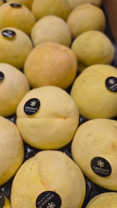 The image shows a close-up, overhead view of a tray of yellow peaches. The peaches are round and have a slightly fuzzy texture. Several of the peaches have black circular stickers with white text that reads "icepeach" and a snowflake graphic, along with the words "sweet, cool & tasty". The peaches are arranged in rows within a dark tray. The lighting is bright and evenly distributed, suggesting an indoor setting, likely a market or grocery store. The background is blurred, focusing attention on the fruit. There are no people or animals visible in the image. The scene is devoid of any recognizable landmarks or elements that would associate it with Līvāni, Latvia.