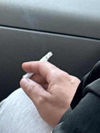 The image shows a close-up of a person's hand holding a cigarette. The hand is light-skinned and appears to be in the process of smoking the cigarette, as evidenced by the way it is held between the thumb and forefinger. The cigarette is white with a brown tip, suggesting it is lit. The background is dark and textured, possibly a piece of clothing or furniture. To the left of the hand, there is a lighter-colored, vertical element with a distinct line, which might be part of the setting. The overall lighting suggests an indoor or shaded environment, and there are no clear indicators of time of day or weather conditions. The image is a close-up, focusing solely on the hand and the cigarette, and does not reveal the full scene, location, or any emotions. There is no visible text in the image.