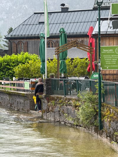 A person wearing a brown hooded jacket and black pants stands on a stone walkway next to a body of water. They are holding a yellow object in their left hand. A child in a green garment stands near their feet. The stone walkway is bordered by a green metal fence. Behind the fence are tables and chairs, likely part of an outdoor seating area for a cafe. In the background, a wooden building with a dark metal roof is visible, with several green and red umbrellas covering parts of the outdoor seating area. A sign on a pole reads "Seecafé Hallstatt - Salzkammergut" and lists food and drink options. The scene appears to be set in a town, possibly with a canal or river, under overcast skies. The environment suggests a temperate climate.