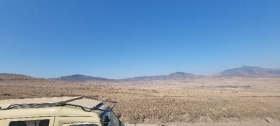 The scene depicts an expansive, arid landscape in the Malanja Depression, Tanzania, under a clear, bright blue sky. In the lower-left foreground, the upper section of a light-colored utility vehicle is visible, equipped with a black, slatted roof rack. The ground consists of vast, flat to gently rolling terrain covered with sparse, dry, light brown and tan vegetation. A lengthy chain of distant, dark, undulating hills and mountains forms the horizon line. No individuals are present, and no specific actions or interactions are depicted.