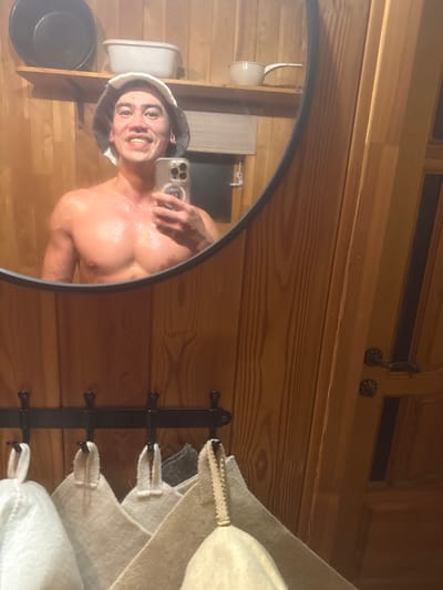 A man wearing a sauna hat takes a selfie in a round mirror. The mirror reflects his face, bare chest, and the hand holding a phone. The background shows the wooden interior of a sauna, including shelves with buckets, a metal hook rack with white felt hats, and wooden paneling. The image is rotated to the side.