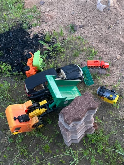 An outdoor, high-angle, eye-level shot shows several overturned toy trucks scattered on a patch of dirt, grass, and sand. In the upper left corner, a pile of black, burnt debris sits amongst green grass and weeds. To the right of the debris, an orange and green toy truck is lying on its side. A black toy SUV with a green dump bed is in the center of the frame. To the right of the SUV, an orange toy tanker truck sits on top of a grey rock. Below the tanker truck, a yellow toy truck with a grey chassis is overturned. In the lower right of the frame, a stack of interlocking paving stones sits in the dirt. The overall impression is of a disused, neglected area.