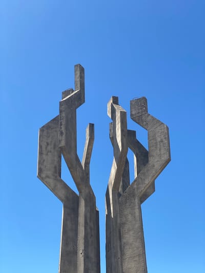 A low-angle shot depicts a concrete architectural structure against a clear, bright blue sky. The structure comprises multiple interconnected, angular beams, giving it an abstract, skeletal appearance. The concrete has a raw, unfinished texture. This is a memorial park in the city of Barutana, Montenegro.