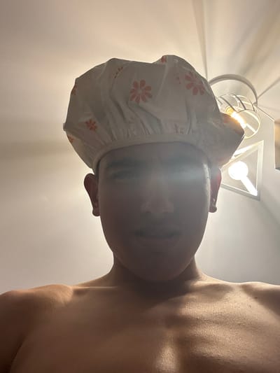 The image depicts a low-angle, head-and-shoulders portrait of an individual from the upper chest upwards. The person, appearing to be an adult male, is looking directly at the camera. They are wearing a white shower cap adorned with orange floral patterns. Their face is largely in shadow, with a bright glare across the eye level, obscuring specific facial features. The individual's shoulders and upper chest are bare. In the upper right background, two distinct light fixtures are visible against a light-colored ceiling. One fixture is a spherical or caged design emitting warm light from a visible bulb. The other, positioned to its right and slightly lower, is a white, angular, possibly triangular fixture with a glowing internal element. The environment is indoors. No visual elements within the image provide information to identify the specific location as Kanchevo, Bulgaria.