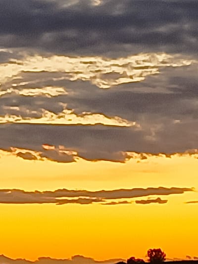The image captures a sunset over a rural landscape. The sky is a gradient of warm colors, transitioning from a deep orange at the bottom to lighter yellow and then to gray and dark blue clouds at the top. Horizontal cloud formations are visible, some illuminated by the setting sun, creating streaks of bright yellow and white. In the foreground, a dark silhouette of the landscape is visible, with hints of trees and possibly low-lying structures. The image is abstract and lacks specific details of buildings or infrastructure that would definitively place it within Somaglia, Italy, but the style of the rural silhouette is consistent with the Italian countryside.