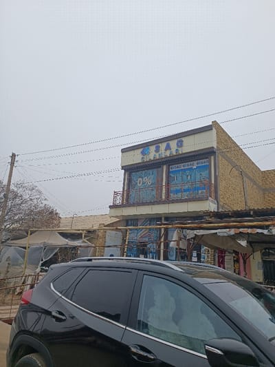 A black SUV is parked on a street. In the background, a two-story building with a sign that reads "SAG GILANLARI" is visible. The upper level has a balcony, and the ground level is a storefront. Other buildings and temporary market stalls with canopies can also be seen along the street. A utility pole is visible on the left side. The sky is overcast.