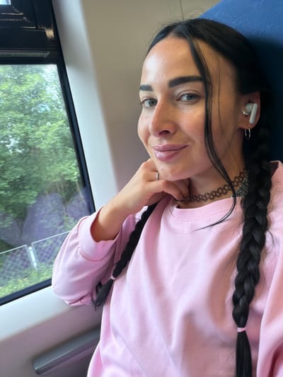 A young woman with dark hair styled in two braids is captured in a selfie from the chest up, seated on a train. She is wearing a pink sweatshirt, a black choker necklace, and white wireless earbuds with a diamond-studded accessory. Her left hand rests on her chin, and she wears a silver ring on her left index finger. A subtle smile plays on her lips, and her eyes are looking towards the camera.
The background reveals a train window on the left, through which lush green trees and foliage are visible, suggesting a journey through a rural or suburban area. The window itself has a dotted pattern. To the right of the woman is the blue upholstered seat of the train.
This scene, with its train interior and a glimpse of passing greenery, could plausibly be observed in many locations. However, if this were to be specifically located within the city of Ушаки, Russia, it would represent a common mode of transportation for residents or visitors traveling within or near the region. The presence of trees outside the window is consistent with the natural landscapes found around Ушаки.