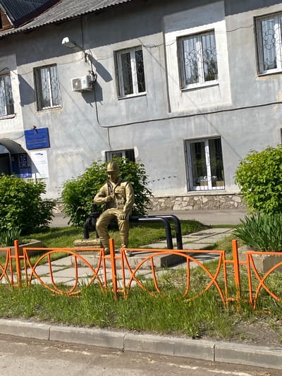This image captures a street scene in Usolye-Sibirskoye, Russia, on a sunny day, likely mid-day or early afternoon, as evidenced by the distinct shadows.
The main subject is a multi-story building with a light gray or off-white stucco facade. It features multiple rectangular windows, each secured with decorative black metal grates. An air conditioning unit is mounted on the wall of the building, and various electrical cables run along its exterior. On the lower part of the building, a prominent blue sign is visible, displaying text in Cyrillic. The most clearly discernible word on the sign is "ПОЛИЦИЯ" (Police), indicating the building is likely a police station or a related administrative office. Additional text below suggests office hours or departmental information.
In front of the building, nestled within green bushes, stands a golden-tan statue of a person. The figure, dressed in what appears to be a uniform and helmet, is depicted in a crouching or kneeling position, holding what resembles a rifle or similar equipment. The statue adds a commemorative or thematic element to the scene.
An orange metal fence with decorative, intertwined circular patterns separates the building's immediate surroundings from the street. This fence runs alongside a paved area, a strip of green grass, a concrete curb, and an asphalt road. The setting combines elements of urban architecture with natural greenery and public infrastructure.