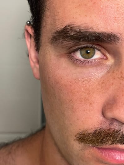 The image is a close-up of a man's face. He has short, dark hair and a light complexion with freckles. He has a piercing in his left ear, and a brown mustache. His right eye is visible and is green in color. The photo was taken indoors against a plain, light background. 

The coordinates provided correspond to a location in Alon HaGalil, Israel. However, it's impossible to know the exact location based on the image.