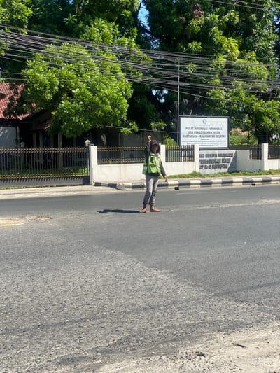 A single individual, wearing a high-visibility green vest and grey pants, stands in the middle of a paved road. The individual is barefoot and appears to be signaling with their left hand, which is raised. A large tree with lush green foliage dominates the background, with power lines strung across it. To the right of the tree, a white sign is visible, bearing text in Indonesian. The sign appears to be an information center, with the words "PUSAT INFORMASI PARIWISATA DAN PENGGOSOKAN INTAN MARTAPURA - KALIMANTAN SELATAN" visible. Behind the sign and the tree, a low-rise building with a tiled roof and a black metal fence is partially visible. The road surface is asphalt with visible tire marks and some debris. The scene appears to be in a tropical or subtropical climate, given the dense foliage and bright sunlight. The location is identified as Sungai Paring, Indonesia.