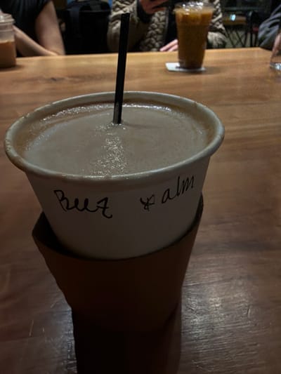 A paper cup filled with a light-colored beverage sits on a wooden surface. The cup has a brown cardboard sleeve. A black straw extends into the drink. Handwritten on the cup is the phrase "Buzz & calm." Another beverage, served in a glass, sits to the left, slightly out of focus. An arm holding the cup is visible, along with portions of other people and furniture in the background.