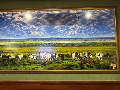 The image displays a framed painting on a green textured wall. The painting depicts a vast grassy plain under a dramatic sky filled with scattered white clouds and blue patches. In the middle ground, a herd of cattle, mostly light-colored with some darker individuals, grazes in the tall grass. To the right, a person on horseback, wearing a hat, blue jeans, and a plaid shirt, appears to be herding the cattle. The horse is brown. In the far background, a treeline separates the plain from a body of water or a fertile valley. In the lower right corner of the painting, a silhouette of Venezuela is superimposed with the text "Llanos Venezuela". This painting is located in the restaurant "El Mesón de Torreluz" in Torrejón de Ardoz, Spain.