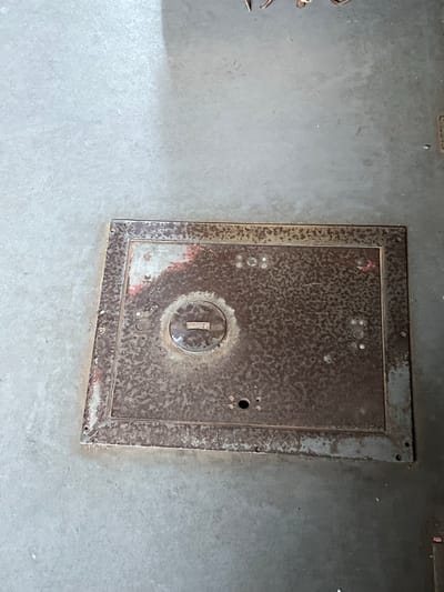 A rusted metal manhole cover is situated on a concrete surface in Sviatopetrivske, Ukraine. The cover is rectangular with a slightly raised, textured surface that displays significant signs of corrosion, including pitting and reddish-brown discoloration. It is bordered by a weathered metal frame that appears to be bolted to the surrounding concrete. A circular indentation with a slot is present near the center of the cover, suggesting it may be a locking mechanism or access point. The concrete surface is a uniform light gray and shows minor scuffs and debris.