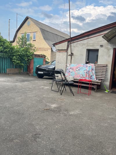 The image depicts an outdoor scene at what appears to be a residential or commercial property in Nemishaieve, Ukraine. In the foreground, a gray asphalt surface is visible, with some scattered debris such as dried leaves and twigs. To the right, a green mop leans against a wooden structure.
In the mid-ground, a weathered wooden bench is positioned against a light gray wall. A colorful blanket with a cartoonish animal pattern is draped over the bench. In front of the bench, a small, red, table with thin legs stands on the asphalt. Two black folding chairs are placed near the table.
Further back, a black SUV with a license plate reading "KA 4332 MC" is parked to the left. Behind the car, a teal-colored metal gate is visible, along with a red brick wall. Lush green foliage grows on a wooden fence to the left of the gate.
In the background, a two-story building with a dark gray, pitched roof constructed of what appears to be slate tiles dominates the left side of the frame. The building has light yellow brick walls and features several windows. A tall, slender metal pole with an antenna-like structure extends upwards from the left side of the building. The sky above is partially cloudy, with white and gray clouds against a pale blue backdrop. The overall scene suggests an unkempt, utilitarian outdoor space.