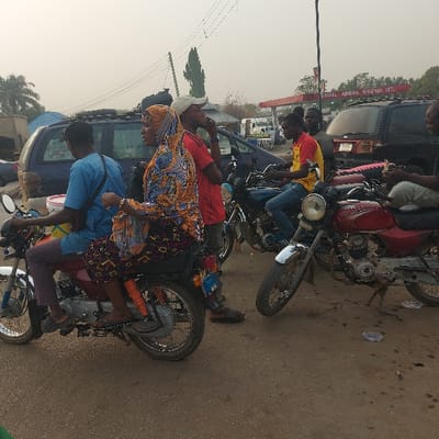 A crowded street scene shows multiple motorcycles parked closely together. People are sitting on some of the bikes, others stand nearby. The riders and passengers are casually dressed. A building with the sign "ISMAIL ABBAS NIGERIA LTD." is visible in the background. A variety of vehicles, including a van and a truck, add to the crowded nature of the scene. The overall impression is one of urban activity and casual transportation.