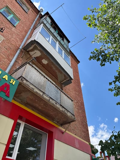 The image shows a close-up, worm's-eye view of the exterior of a brick apartment building, likely in Kamensk-Shakhtinsky, Russia. The building has several balconies, one of which is enclosed with white siding and windows, and another below it with corrugated metal siding. A large green sign with yellow and red lettering is visible in the lower left corner, partially obscuring a red and yellow storefront. A tall, thin metal pipe runs vertically along the brick wall. Above the building, a clear blue sky with scattered white clouds is visible, and the leaves of a green tree overhang the scene from the right. The overall impression is one of urban architecture under a bright, sunny sky.