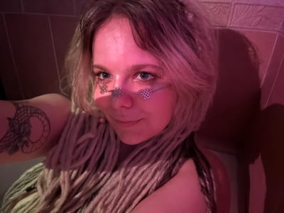 A woman with light-colored dreadlocks and light-colored hair is shown in a selfie. She is wearing a silver chain across her nose and cheeks. She has a tattoo on her left arm that appears to be a stylized dragon. The background suggests she is in a tiled shower or bathroom, with textured tiles visible. The lighting is dim and pinkish, possibly from a colored light source. There is a shadow cast on the wall behind her. The image appears to have been taken in a domestic setting within the city of Vostochny District, Russia.