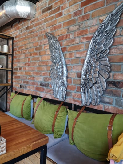 The image depicts an interior scene, possibly a cafe or restaurant, featuring a brick wall adorned with two large, metallic angel wings. In the foreground, a wooden table with a black metal frame is visible on the left. Behind the table, a bench seating area is furnished with several large, olive-green cushions secured with brown leather straps. The wall is made of red brick with visible mortar. A silver ventilation duct runs across the upper left portion of the wall. A black metal shelving unit with two shelves, holding two white candles and other decorative items, stands to the left of the wings. The overall aesthetic suggests a modern, industrial-chic design. There are no people visible in the image. The provided crops focus on specific details of the brick wall, the angel wings, the cushions, and the table. The location provided, Yantarny, Russia, is context for the image but not directly depicted within it.