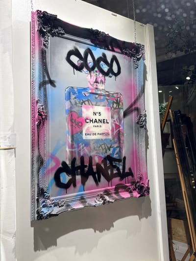 A close-up shot shows a framed piece of art hung on a white wall. The artwork features a Chanel No. 5 perfume bottle design with graffiti-style elements, including spray-painted words and symbols in black. The frame is ornate, with a silver-gray and black finish, and touches of pink and blue. The art is labeled as "Ghost Coco Chanel Graffiti, Limited Edition of 23, €1695". The surrounding environment appears to be a store or gallery, as indicated by the retail price tag and the glimpse of other framed items on a shelf in the upper background, also there are plants in the background. The location of the image is Windermere, United Kingdom.