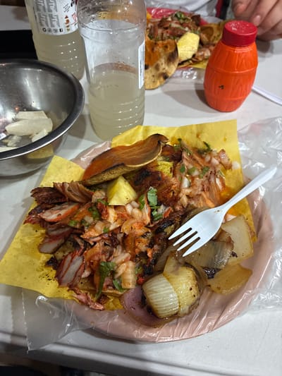 An overhead, medium shot captures a plate of al pastor tacos, served on yellow paper, on a light-colored table. The tacos are made with thinly sliced, marinated pork, garnished with chopped white onions and cilantro, and topped with a slice of pineapple. A white plastic fork rests on the plate. In the background, two plastic bottles of a pale yellow liquid, likely a beverage, are visible. To the right, a small, orange plastic squeeze bottle with a red cap, likely a condiment dispenser, sits on the table. The table surface is partially covered with a clear plastic bag, which also drapes over the plate. The overall setting appears to be a casual dining environment, possibly a street food stall in Tijuana, Mexico, given the style of the food and common accompaniments.