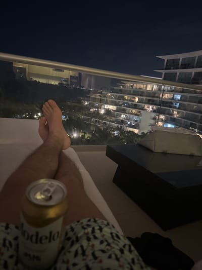 At night, a man reclines on a balcony, holding a Modelo beer can. His legs, crossed at the ankles, are visible. A white towel or blanket is beneath him. In the background, a multi-story building, possibly a hotel or apartment complex, is illuminated with many lights. Trees can be seen between the building and the balcony. The balcony features a glass railing and dark furniture. The sky is a dark blue.