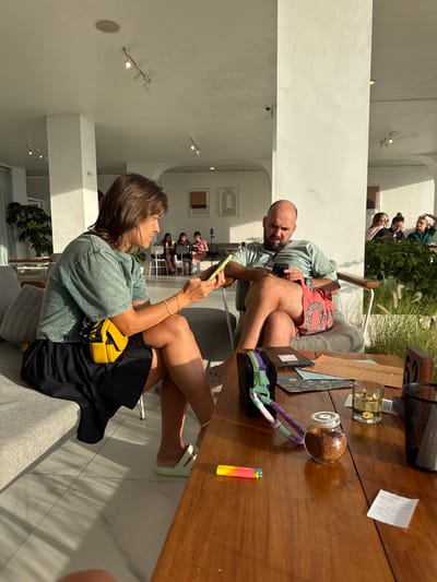 The scene is an interior setting, likely a cafe or restaurant, located in Kedisan, Indonesia. The image features a light-skinned man and a woman sitting at a wooden table. The man is wearing a light green t-shirt and red patterned shorts. He is holding a cell phone and looking towards the camera. The woman, seated facing him, also holds a phone and is wearing a light green t-shirt, a black skirt, and a yellow purse. 

On the table, there is a candle, a small glass jar, a lighter, menus, and a black pencil case. In the background, there is a group of four people standing in the doorway. The cafe/restaurant appears modern with white walls and arched doorways. The environment seems bright and airy.