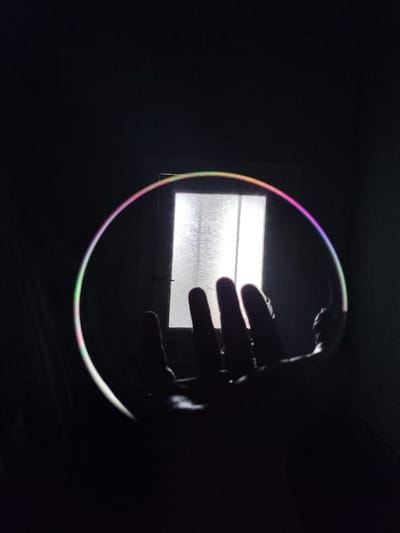 A hand is holding a bubble in front of a window in a dark room. The bubble reflects the light from the window and displays iridescent colors. The window appears to be a paneled door with frosted glass. The overall scene is dark, with the bubble and window being the primary sources of light.