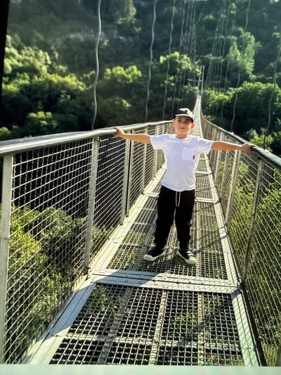 A young boy stands on a metal mesh suspension bridge with his arms outstretched, leaning on the railing. He is wearing a white t-shirt, black sweatpants, a baseball cap, and dark sneakers. The bridge is surrounded by lush green trees and foliage, indicating an outdoor, natural environment, likely a forest or mountainous region. The bridge itself appears to be part of a park or recreational area. The sky is visible above, suggesting it is daytime.