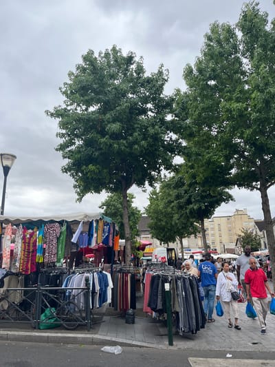 This is a street-level, eye-level, medium shot capturing a scene at a market in Villeparisis, France. The image is dominated by a stall selling clothing, with racks of garments displayed on the left and right. Several people are browsing or walking through the market.

In the foreground, a street market is in full swing. On the left, a clothing stall features a variety of colorful dresses and tops hanging on display. Below these, racks of trousers and other apparel are neatly arranged. A bicycle with a green bag is leaning against a metal fence in front of the stall. The ground is paved with grey cobblestones and asphalt.

In the midground, more clothing stalls are visible, with an assortment of shirts, jackets, and other garments. Several individuals are walking along the market path. A woman in a white top and light-colored trousers is walking towards the right, carrying blue shopping bags. Beside her, a man in a red polo shirt and light-colored trousers is also walking to the right, carrying blue shopping bags. Behind them, a man in a grey t-shirt and a woman in a white headscarf are browsing the stalls. Further back, a van and other market stalls are visible.

In the background, tall green trees with lush foliage are prominent, partially obscuring buildings. The buildings appear to be apartment blocks with light-colored facades and numerous windows. The sky is overcast and grey. The overall impression is a lively outdoor market on a cloudy day.