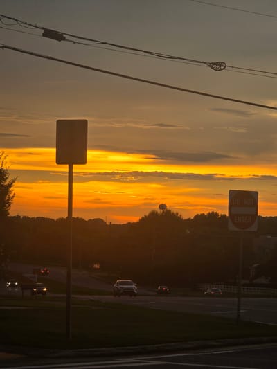 An evening view of a street in Woodstock, United States of America. The sky is a vibrant mix of orange and yellow due to the setting sun, with scattered clouds. In the foreground, a street sign with a blank rectangular plate on a pole is visible, alongside a "DO NOT ENTER" sign. The street is visible with traffic moving, including a white SUV and other cars. Power lines stretch across the top of the frame. In the background, a water tower stands on a hill, surrounded by trees. The overall scene depicts a typical suburban or semi-urban environment at dusk.