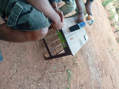 Two people are sitting outside, one wearing denim shorts and the other khaki pants. A small wooden table separates them, holding a smartphone, a water bottle, and another electronic device. The table has a green and white design on its side. The ground is a mix of dirt and dry grass. The scene has a casual, outdoor feel.