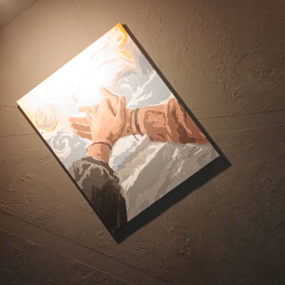 A painting is displayed on a textured, dark brown wall. The painting depicts two hands clasped together, rendered in a stylized, somewhat abstract manner with flat areas of color. The hands are depicted in shades of tan and brown, with darker brown bracelets or bands on the wrists. Surrounding the hands are abstract shapes in white, grey, and light orange, possibly representing clouds or swirling energy. The painting is lit by a spotlight from the upper left, casting a shadow on the wall to the right and below the artwork. The overall impression is of a piece of art displayed in an indoor setting. The location is Vyshgorodok, Russia.