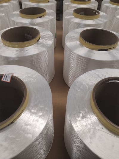 This media file shows several large spools of white, translucent material, likely fiberglass yarn or thread, arranged in rows. The spools are cylindrical with cardboard cores and yellow tape around the edges. The material on the spools is wound tightly, creating a textured, slightly shimmering surface. The lighting suggests an indoor setting, possibly a warehouse or factory, with light coming from above and to the right, creating highlights and shadows that emphasize the texture of the material. The arrangement of the spools indicates a manufacturing or storage environment. Some of the spools have labels attached to their cores, though the text on these labels is mostly illegible. One label is partially visible and appears to have alphanumeric characters and possibly some Dutch text, given the location context of Deurne, Netherlands. The overall impression is one of industrial production and supply. No people are visible in the image, and the scene is static. The time of day and weather are not discernible from the image.