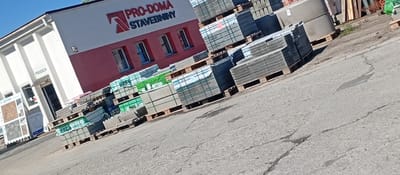 The image displays the exterior of a building supply store, "PRO-DOMA STAVEBNINY," located in Karviná, Czechia. The primary structure is a commercial building with a white upper facade and a reddish-brown lower facade. The store's name, "PRO-DOMA STAVEBNINY," is prominently displayed in red and white lettering on the reddish-brown section, accompanied by a red, arrow-like logo. Below this signage, multiple windows with visible security bars are present. On the white section of the building, an entrance is marked by the word "PRODEJNA" (SHOP) above it. To the immediate left of the entrance, an outdoor display stand showcases samples of paving or roofing materials, with partial visibility of the brand "DITON."

The foreground and the area immediately adjacent to the building are occupied by numerous wooden pallets stacked with grey concrete blocks or paving stones. Many of these stacks are wrapped in either green or clear plastic, with some green wraps showing partial text suchs as "ORES" and "BETON OL." Further in the background, large cylindrical concrete structures, possibly for wells or drainage, are also stacked on pallets. Other construction materials, including lumber and metal shelving units, are partially visible in the distance. The ground consists of asphalt pavement, characterized by multiple visible cracks and signs of wear. The scene is brightly illuminated by direct sunlight, casting some shadows. No individuals are present in the image.