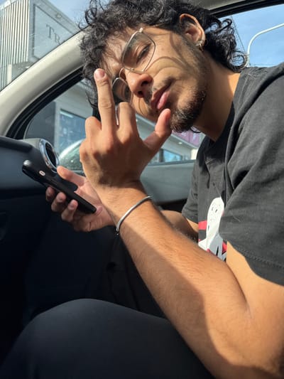 The scene is located within the city of Tolhuin, Argentina. A male individual, appearing to be a young adult, is seated in the passenger compartment of a vehicle during daylight hours. He has dark, curly hair, a short beard, and wears glasses with thin, light-colored frames. His attire consists of a black short-sleeved t-shirt with a white graphic print and dark trousers. A plain silver-colored bangle bracelet is on his right wrist. His left hand is raised to face level, with the thumb, index, and little fingers extended, and the middle and ring fingers curled. His right hand holds a black smartphone. Strong, directional sunlight casts prominent shadows across his face, arm, and chest. The vehicle's dark interior includes a visible air vent with a metallic ring. Through the vehicle's window, a blurry exterior background shows parts of a building with reflective or metallic vertical elements and a sign partially visible, displaying the letters "TR". The sky is clear and blue.