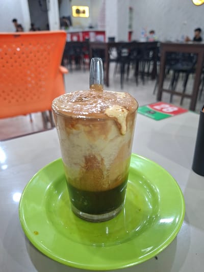 A glass of iced coffee with milk and a green base is placed on a bright green saucer on a white table. The drink has a foamy, light brown top layer, with streaks of white and brown within. The green base is visible at the bottom of the glass. In the background, the blurry interior of what appears to be a cafe or restaurant is visible. Orange chairs and dark wooden tables with chairs are present. Faint figures of people can be seen in the background. A red and green floor mat with the number "28" is visible in the middle ground. The overall lighting suggests an indoor environment, possibly during the daytime. This scene is likely located in the city of Sigli, Indonesia, given the context provided.