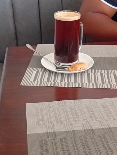 The image depicts an indoor scene, characteristic of a cafe or restaurant setting, in Murang'a, Kenya. A polished dark brown wooden table surface dominates the foreground. Two woven placemats are visible; the upper one features a pattern of alternating light grey and dark grey vertical stripes, while the lower is solid light grey with a textured weave. A white circular saucer rests on the upper placemat, holding a tall, clear glass mug filled with a dark, reddish-brown beverage topped with a thin layer of light foam. A silver spoon lies diagonally on the saucer. Adjacent to the mug, a single, oval-shaped, light brown cookie is present on the saucer. In the background, a dark grey upholstered booth seat with vertical channel tufting is visible on the left. On the right, the upper arm and shoulder of an individual with brown skin are partially seen, wearing a garment with a white and dark-colored stripe.