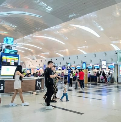The image captures a bustling scene inside a modern airport terminal in Sidourip, Indonesia, likely during the daytime. The expansive space features a high, arched white ceiling with recessed linear lighting, contributing to a bright and contemporary atmosphere. The floor is tiled with a light color, incorporating dark rectangular patterns that delineate pathways.
In the foreground, several individuals are walking or standing. A woman in a white two-piece outfit and white sneakers walks towards the left. Near the center, a man in a black t-shirt and dark pants, along with another adult, observes a young child wearing a patterned t-shirt and blue pants, who is walking independently across the floor. Further back, more travelers can be seen moving around the check-in and information areas.
The background is lined with numerous airport check-in counters and self-service kiosks. These counters feature screens displaying various information. On the left side of the image, a prominent blue information stand is visible. It displays a pictogram of two figures with a question mark, indicating an inquiry point. Below this, text reads "Ajuda Rápida / Fast Assistance" and another sign below that says "Péplia Prioritária / Prioritaire" (likely indicating a priority lane). A large screen below these signs shows flight information with colorful bars, possibly denoting status or categories. The walls behind the counters have a distinctive design with circular cutouts. People are engaged in typical airport activities such as queuing, checking in, and navigating the terminal. The overall mood appears calm and orderly.