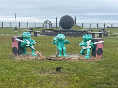 This image depicts an outdoor scene in the city of Kuril'sk, Russia, featuring three turquoise frog statues positioned on a grassy area. Each frog is depicted playing a guitar, and they are accompanied by pink and black speaker-like structures. In the background, a large, dark, spherical sculpture composed of stones is visible, along with a metal archway, a tall monument, and a fence. The sky is overcast and a lawnmower is present in the mid-ground.