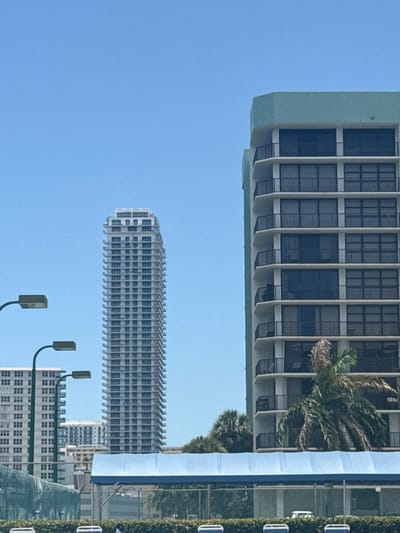 The image captures a daytime urban landscape in Hallandale Beach, United States, under a clear blue sky. A tall, slender, light-colored high-rise building with numerous visible floors, appearing either under construction or recently completed, stands prominently in the center-left. To its right, a multi-story residential building with a light-colored facade and a distinctive light teal/green roof and side accent occupies the right side of the frame, displaying multiple floors with dark-railed balconies. Further to the left and in the background, another light-colored multi-story building is partially visible. The immediate foreground features a blue fabric awning or canopy structure, positioned above a chain-link fence with a green mesh screen attached. Several palm trees and other green foliage are visible behind the fence and awning, interspersed with the buildings. Two dark-colored streetlights with rectangular luminaires curve upwards from the left side of the image. No individuals are visible within the scene.