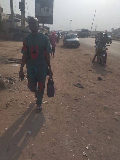 The image shows a man walking on a dirt road in Way, Nigeria. He is wearing a traditional Nigerian outfit with a blue and red pattern, and carrying a black bag. He is walking to the left of the image. There are other people walking in the distance, and a few vehicles on the road. There is a sign in the background. There are broken pieces of glass on the ground. The environment is dusty and dry. The man is in the foreground.
