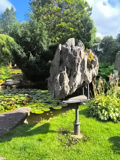 The image shows a garden with a large, uniquely shaped gray rock formation balanced on a metal stand in the foreground. To the left of the rock formation is a pond filled with numerous green lily pads and a few pink lily blossoms. A stone slab edge is visible in the lower left corner of the pond. Behind the pond and rock, lush green trees and foliage dominate the background, with a bright blue sky with scattered clouds visible above. The ground in front of the rock formation is covered in vibrant green grass. The scene appears to be a tranquil outdoor setting, possibly a park or botanical garden. This specific rock formation is a boulder from the Mosėdis boulder park in Lithuania.