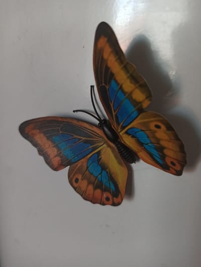 A close-up, top-down shot captures a single, artificial butterfly ornament with orange, blue, and black patterned wings. The butterfly is positioned on a plain, light-colored surface, possibly a wall or table, casting a soft shadow to its right. The background is uniformly lit, with a slight gradient suggesting an overhead light source. There are no people or other discernible objects in the image. The scene is devoid of any context that would place it within the city of Promyshlennaya, Russia, or any other specific location.